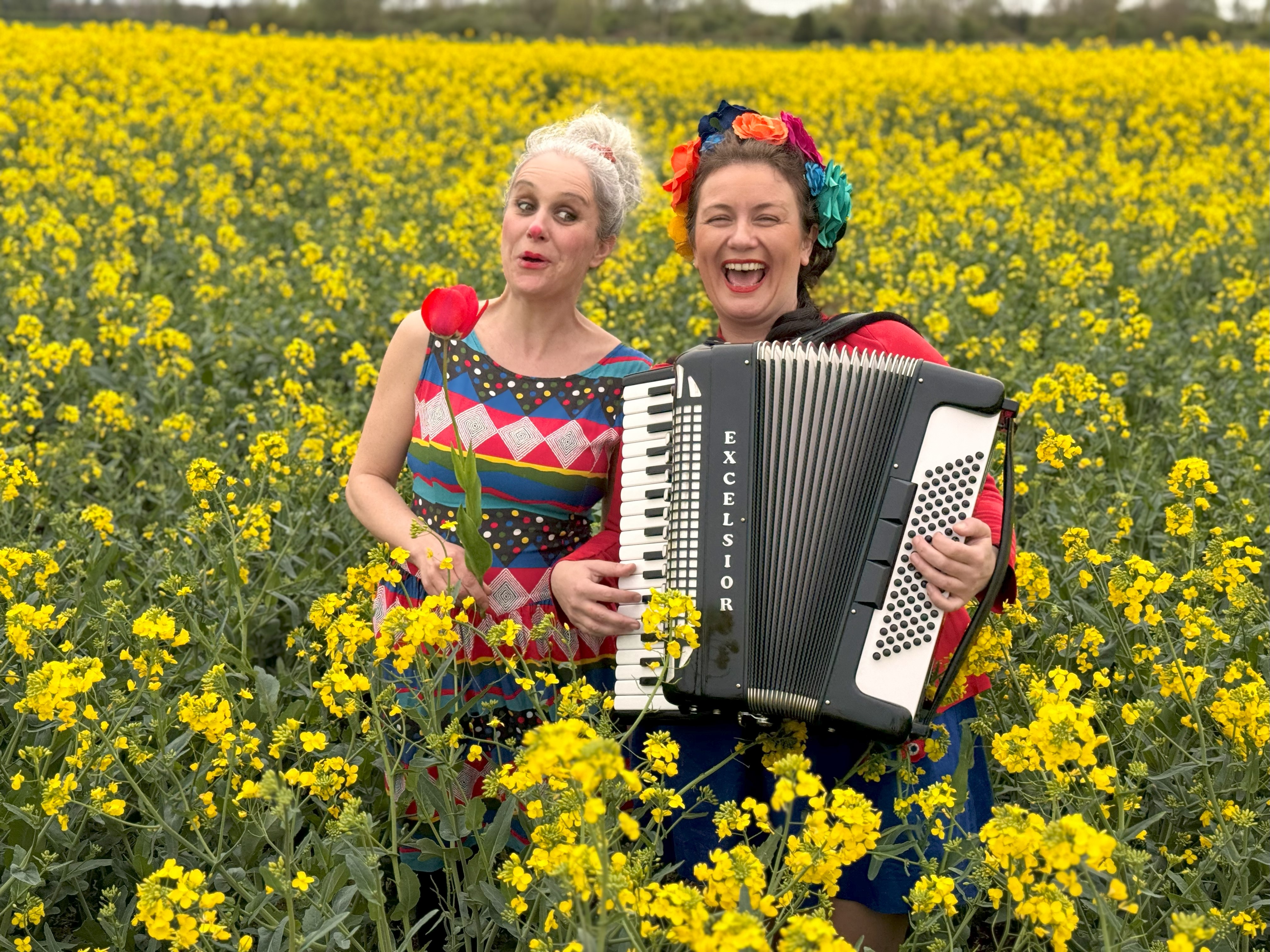 Au milieu d'un champs de colza jaune lumineux, Stephanie Jancar, en robe de clown, tulipe rouge dans la main et Clémence Vandaele chanteuse, fleurs dans les cheveux, jouant de l' accordéon, rayonnent de joie. 