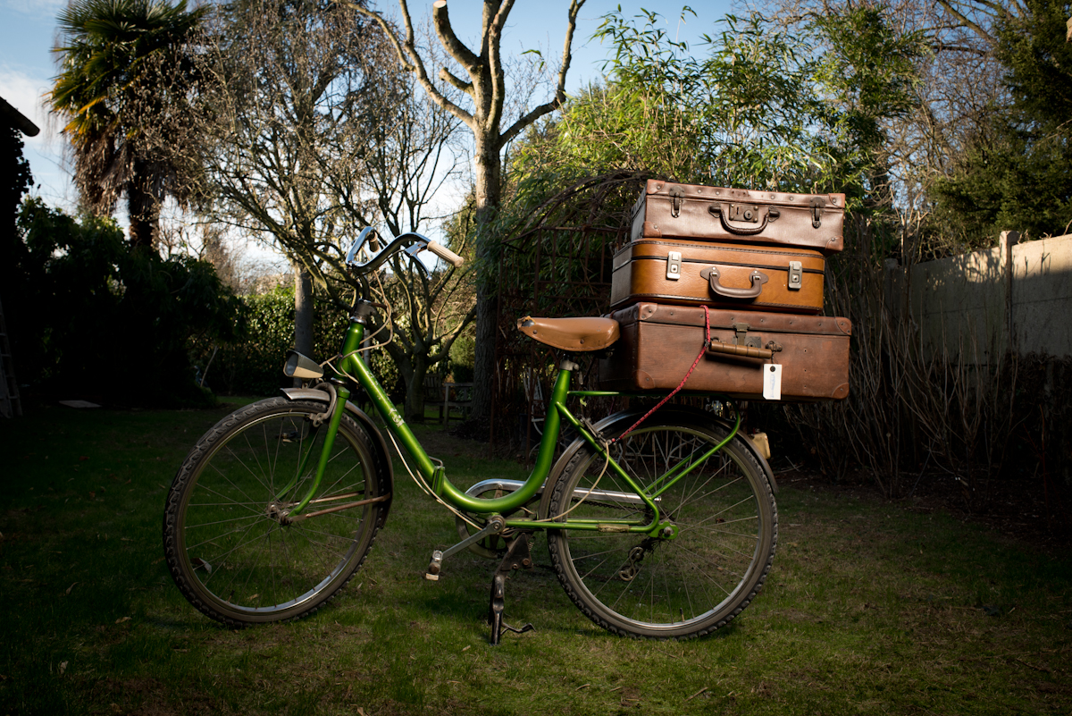 dans un jardin, un vélo vintage vert avec sur son porte bagage, trois valises en cuir marron